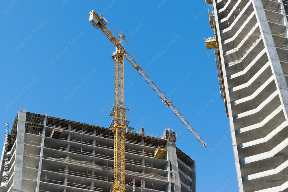 high-rise construction crane with a long arrow of yellow color against the blue sky over a new multi-storey building of concrete and brick under construction