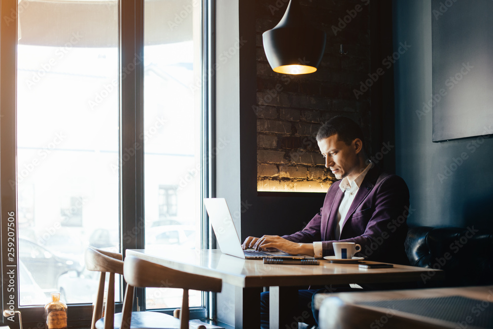Young handsome man sitting in office with cup of coffee and working on project connected with modern cyber technologies. Businessman with notebook trying to keep deadline in digital marketing sphere. Young handsome man sitting in office with cup of coffee and working on project connected with modern cyber technologies. Businessman with notebook trying to keep deadline in digital marketing sphere.