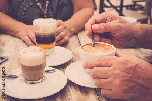 Close-up of hands with coffee cups in a cafe bar enjoying cappuccino for tasty breakfast in the morning - aged hands and mature couple have a break together with drinks on a rustic wooden table