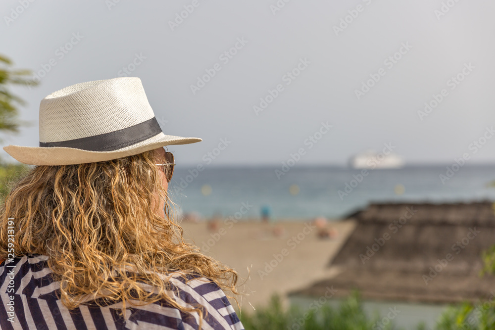 A blond tourist woman with hat enjoys the scenery of a beach on a summer day