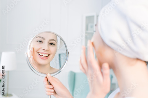 beautiful woman in white bathrobe and towel looking at her perfect skin in mirror and Applying Tonal Cream Base On Face