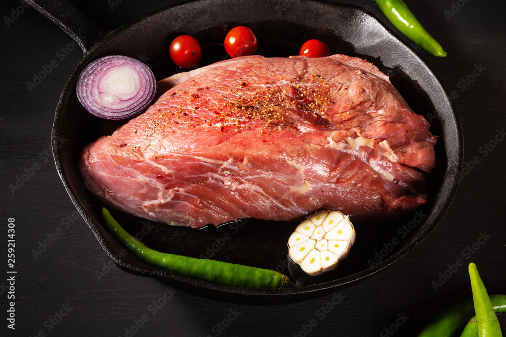 Raw fresh beef in a frying pan and vegetables. Preparation for roast beef. Black wooden table background. Close up. Selective focus.