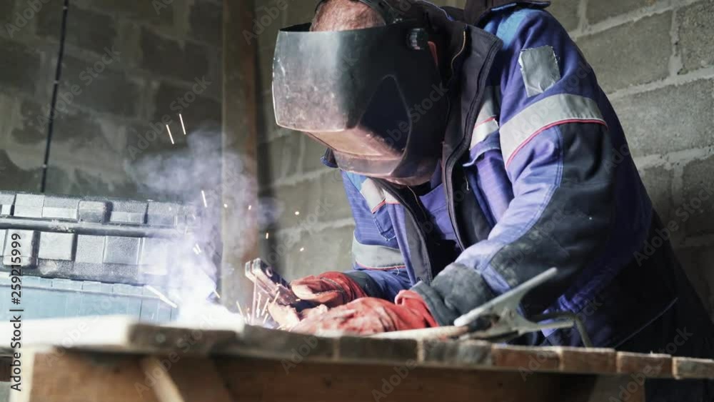 Side view welder in welding mask welds two metal parts. Workman in coveralls is working indoors Side view welder in welding mask welds two metal parts. Workman in coveralls is working indoors