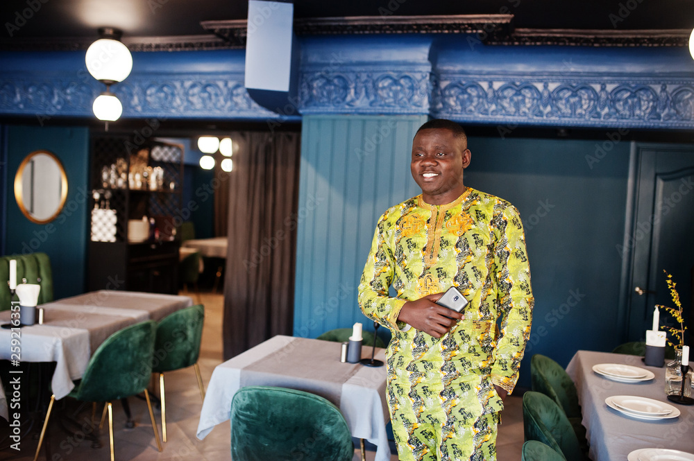 Friendly afro man in traditional yellow clothes at restaurant hold mobile phone at hand. Friendly afro man in traditional yellow clothes at restaurant hold mobile phone at hand.