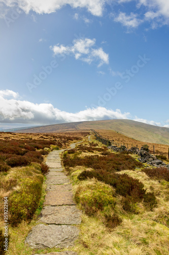 A view of a stony path trail along heater in the hill under a majestic blue sky and white clouds