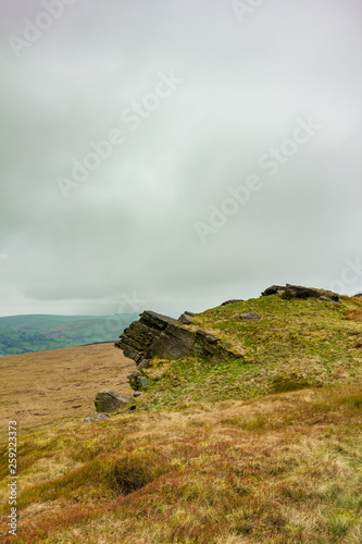 A view of a rocky summit hill with heather and valley in the background under a white cloudy sky