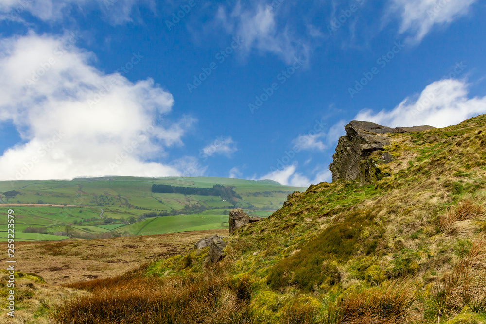A view of a rocky summit hill with heather and green fields valley in the background under a majestic blue sky and white clouds