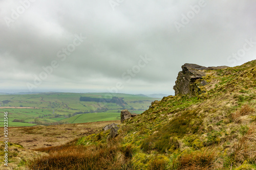 A view of a rocky summit hill with heather and green fields valley in the background under a white cloudy sky
