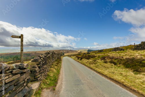 A view of a rural road lane with stone wall on the side  walking path sign and green vegetation under a majestic blue sky and white clouds