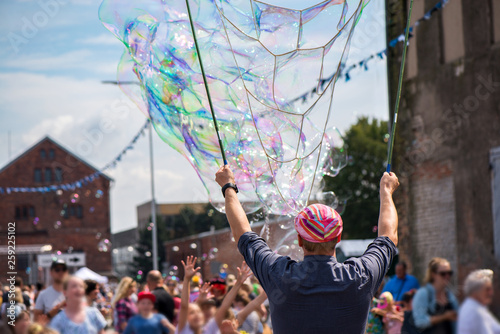 A freelance clown blowing hundreds of tiny, small and big bubbles at outdoor festival in city center. Concept of entertainment, birthdays. Kids having fun. Shower of bubbles flying in the happy crowd
