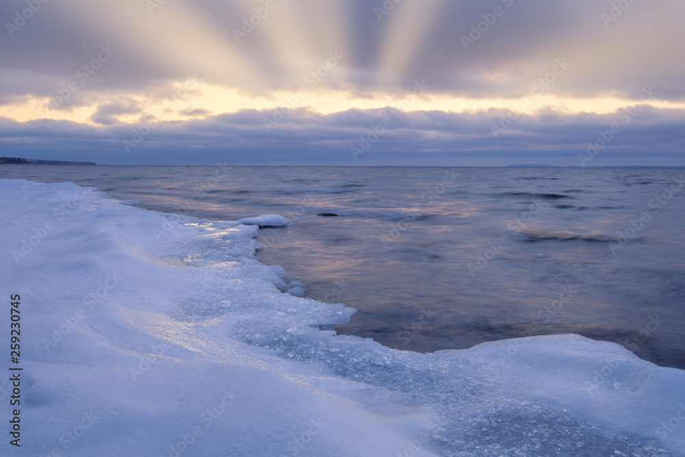 ice and snow on a beach in the foreground, sun rays shining through the clouds in the background