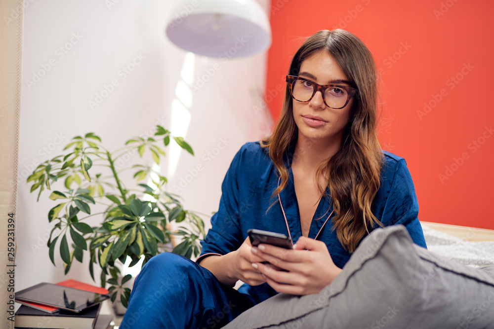 Charming Caucasian brunette dressed in blue pajamas and with eyeglasses looking at camera while sitting on bed in bedrooma and using smart phone. Morning time. Charming Caucasian brunette dressed in blue pajamas and with eyeglasses looking at camera while sitting on bed in bedrooma and using smart phone. Morning time.