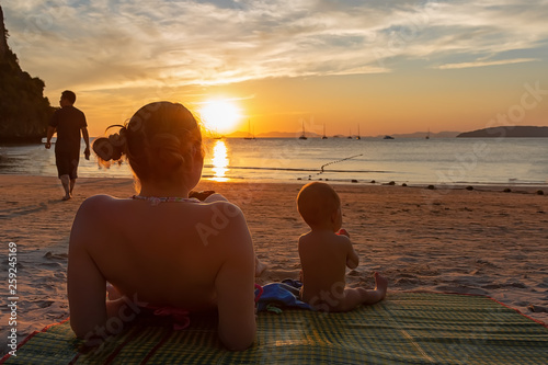 Mother with her daughter. Little baby girl of 9 months. Couple sitting back on a tropical beach  watching and enjoy the sunset. Beautiful blue sky  a bit cloudy  golden sunset light.  Man leaves
