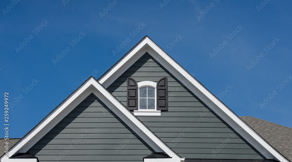 Attic window with black shutter on grey blue siding, gable, corbel, louver on a new construction luxury American single family home in the East Coast USA with blue sky background Attic window with black shutter on grey blue siding, gable, corbel, louver on a new construction luxury American single family home in the East Coast USA with blue sky background