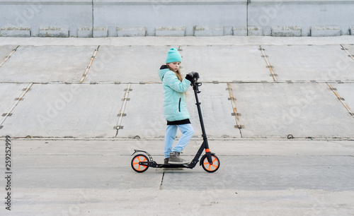 Activities: A school girl with a blue jacket riding a scooter on a gray concrete promenade.