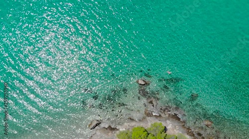 Aerial view of blue sea surface on rocky coast with light reflections. Top view of transparent turquoise ocean water surface and rocks on shore.