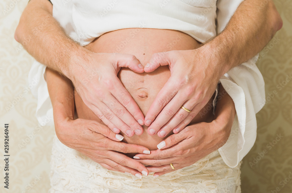 Man holding belly of his pregnant wife making heart. Pregnant woman and loving husband hugging tummy at home. Heart of hands by couple on pregnant belly. Man holding belly of his pregnant wife making heart. Pregnant woman and loving husband hugging tummy at home. Heart of hands by couple on pregnant belly.