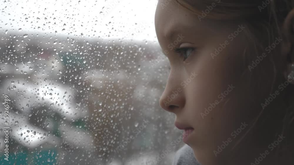 Sad teenager girl looking outside through the window in the rainy day.