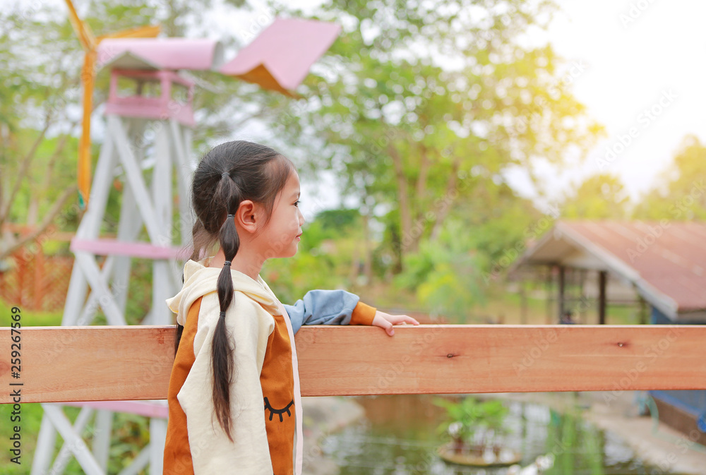Naklejka premium Side view little Asian child girl in warm clothes standing on balcony with looking out in the garden.