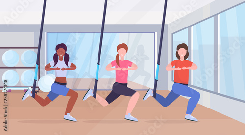 sporty women doing squats exercises with suspension fitness straps elastic rope mix race girls training crossfit group classes workout concept modern gym health club interior full length