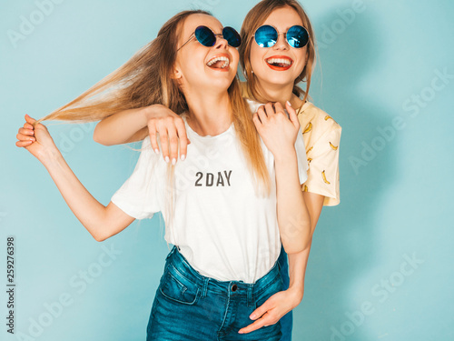 Two young beautiful smiling blond hipster girls in trendy summer colorful T-shirt clothes. Sexy carefree women posing near blue wall in round sunglasses. Positive models having fun © halayalex