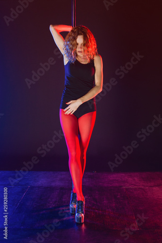 Pole sports: beautiful young athletic fit strong redhead woman gymnast in black T-shirt, shorts and red stockings doing sport exercises (training, work out) on the pole on dark background