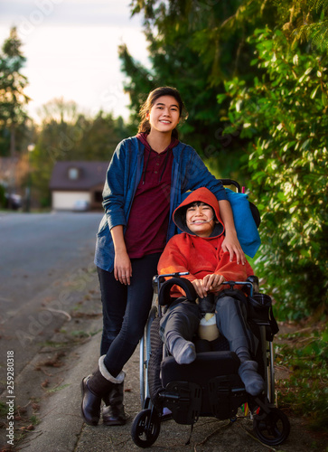 Sister standing next to disabled little brother in wheelchair outdoors