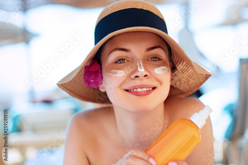 Facial Care. Young  Female Holding Bottle Sun Cream and  Applying on Face Smiling. Beauty Face.  Portrait Of Young Woman in hat Smear  Moisturizing Lotion on Skin. SkinCare