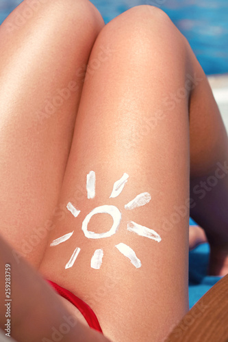 Young woman with sun shape on the leg holding sun cream bottle on the beach. Sun protection sun cream, on her smooth tanned legs. Sunblock. Skincare.