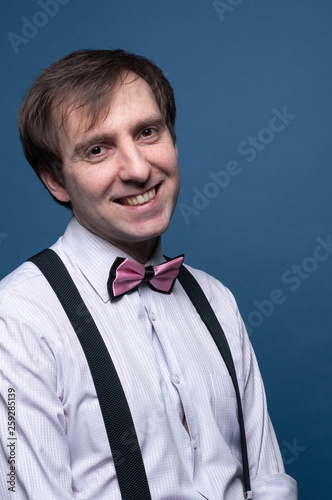 handsome man in shirt, pink bow tie and black suspenders looking at camera and smiling on blue background © Viktoriia