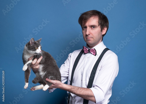 confident handsome man in shirt, pink bow tie and suspender holding on outstretched arms cute grey cat and looking at camera on blue background with copy space