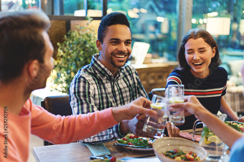 Happy young multi-ethnic friends sitting at table with dishes and toasting at dinner party