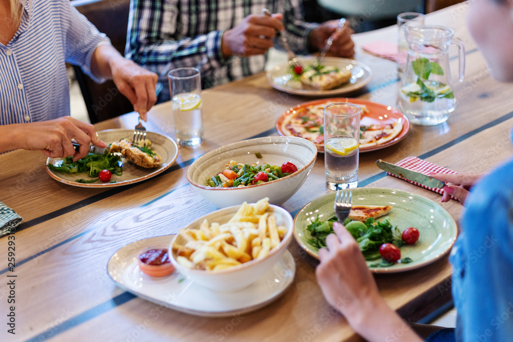 Close-up of main dishes, french fries, vegetable salad and pizza on wooden dining table and three young friends having dinner together Close-up of main dishes, french fries, vegetable salad and pizza on wooden dining table and three young friends having dinner together