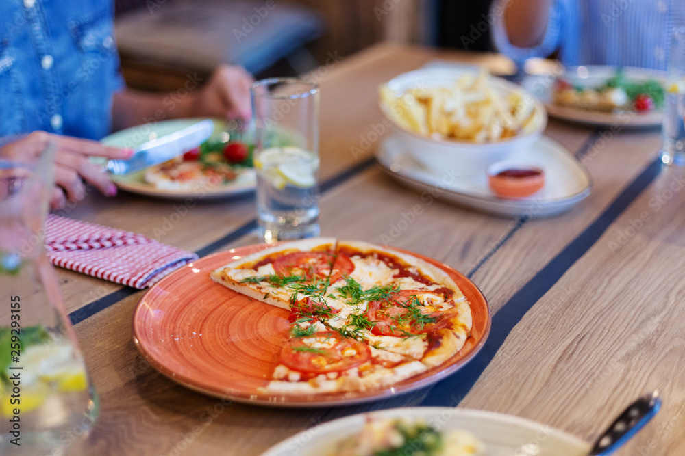 Slices of pizza on red plate on wooden dining table surrounded by other food and drinks and defocused people dining in background Slices of pizza on red plate on wooden dining table surrounded by other food and drinks and defocused people dining in background