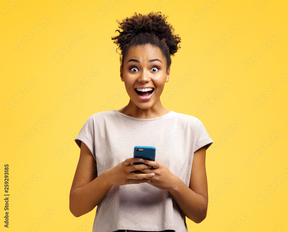 Young girl holds phone, happy to receive notification. Photo of african american girl wears casual outfit on yellow background. Emotions and pleasant feelings concept. Young girl holds phone, happy to receive notification. Photo of african american girl wears casual outfit on yellow background. Emotions and pleasant feelings concept.