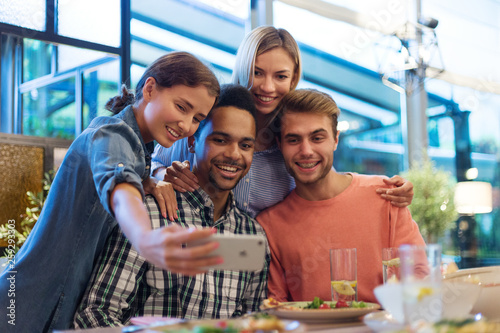 Portrait of four cheerful young friends taking selfie together on smartphone during dinner in cafe