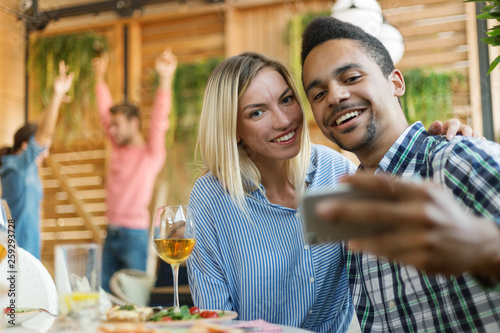 Young woman and man taking selfie together sitting at table at party, their friends having fun dancing in background