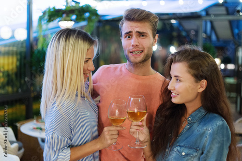 Obraz na płótnie Self-confident young man with dental braces on his teeth posing with two girls clinking wineglasses. Handsome male feeling like womanizer at party w salonie