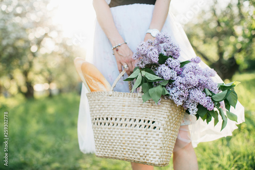 Obraz na plátně Basket with bouquet of lilacs and baguette in woman hands on background of natur