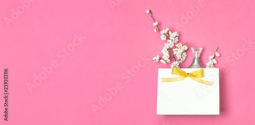 White gift bag with gold ribbon, bottle of perfume, spring white flowers on bright pink background. Greeting card with delicate flowers Pink floral background. Spring minimal concept Flat lay top view photo