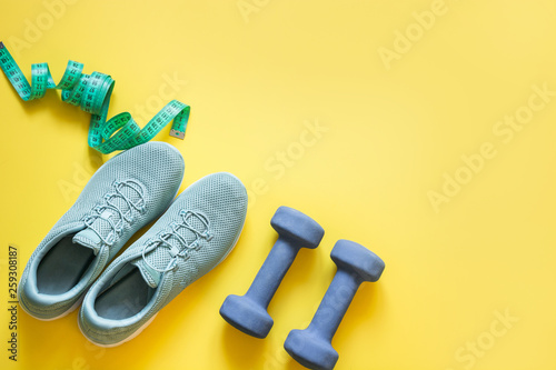Sport and fitness equipment, dumbbells, fitness shoes, measuring tape on punchy yellow. Top view, space for text.