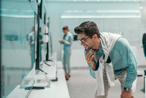 Smiling handsome mixed race man with eyeglasses and dressed smart casual looking at price and specifications of tv he want to buy. Tech store interior.