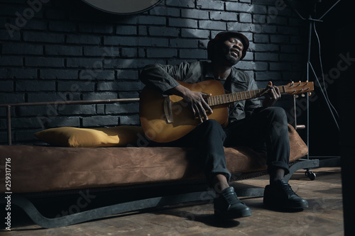 Canvas Print Musician in black trench coat and bucket hat sitting on bed and playing guitar