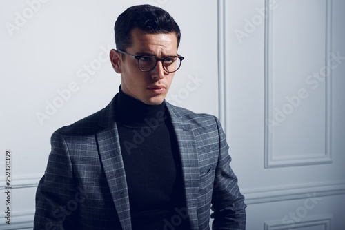 Strong, stylish man in a turtleneck suit,black eyeglasses and gray plaid  jacket looks with attitude at the one side.Horizontal view on the white  wall background. Stock Photo | Adobe Stock