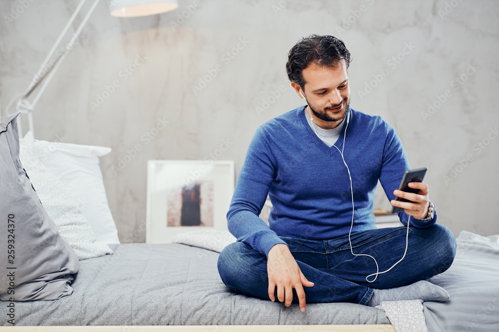 Cute arab man sitting on the bed in bedroom with legs crossed and listening music over smart phone.