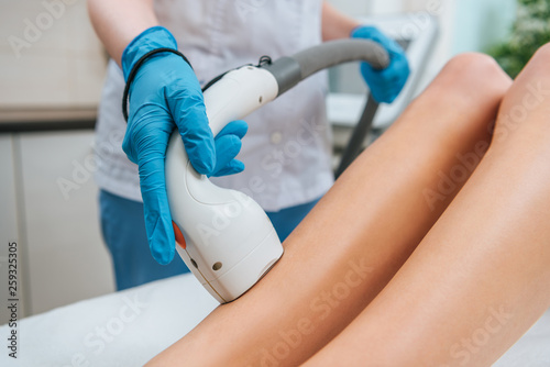 Partial view of cosmetologist in rubber gloves doing laser hair removal procedure on legs