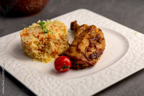 half an appetizing grilled chicken with rice and cherry tomato on white plate, on dark table, horizontal side view