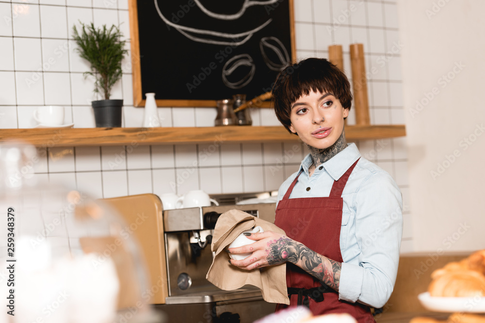 selective focus of pretty barista wiping cup while standing near espresso machine selective focus of pretty barista wiping cup while standing near espresso machine