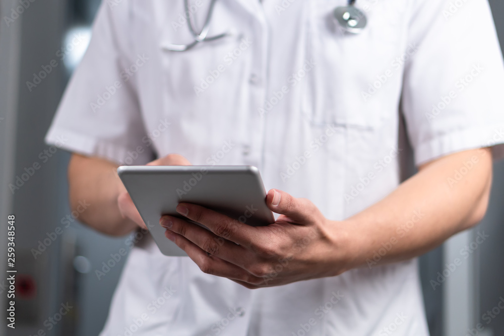 Bright photo of male doctor in uniform with stethoscope coming out of the elevator and using computer tablet in hospital. Modern medical concept Bright photo of male doctor in uniform with stethoscope coming out of the elevator and using computer tablet in hospital. Modern medical concept