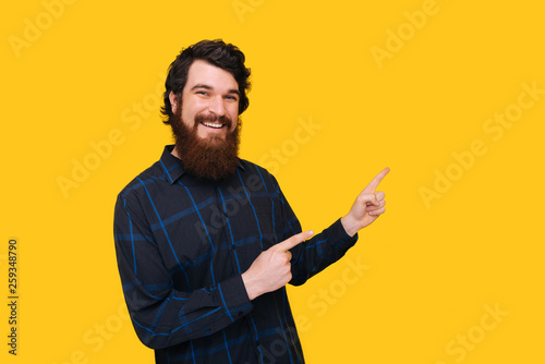Smiling bearded man looking at the camera and pointing aside on yellow background.